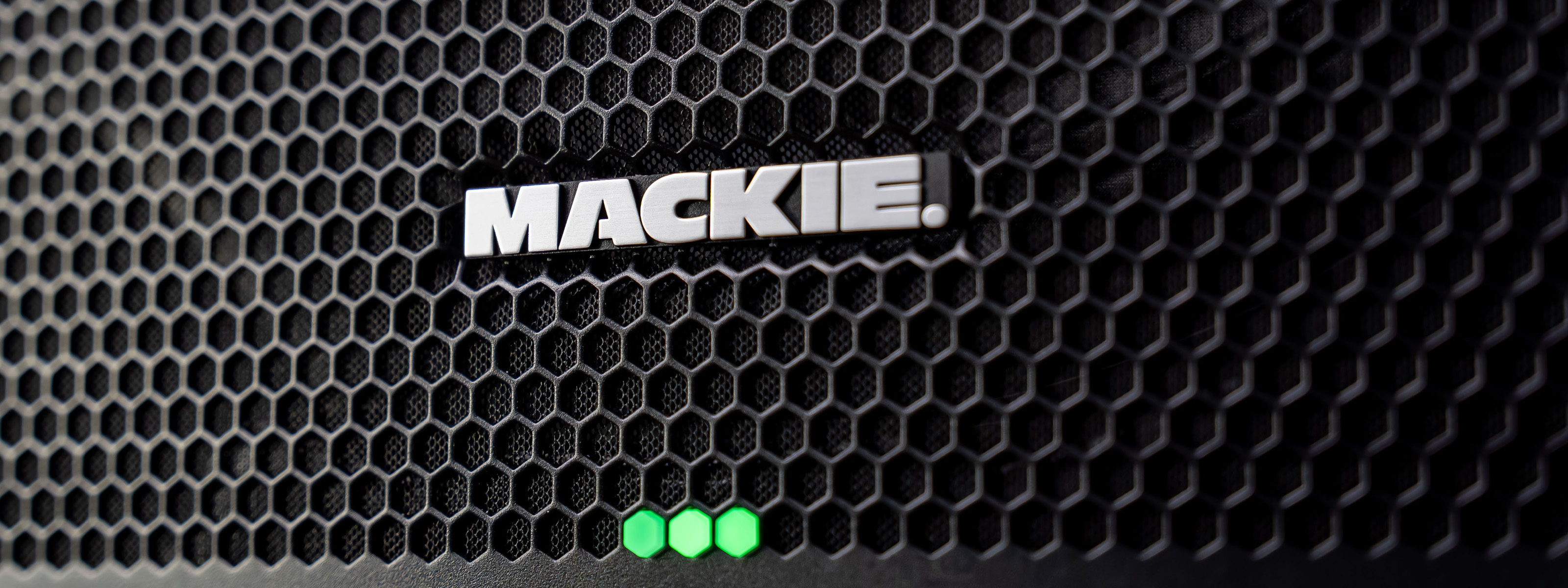 Logotipo da Mackie na parte frontal da caixa de som Thumpv4, cercado por uma grade. Três LEDs verdes abaixo.