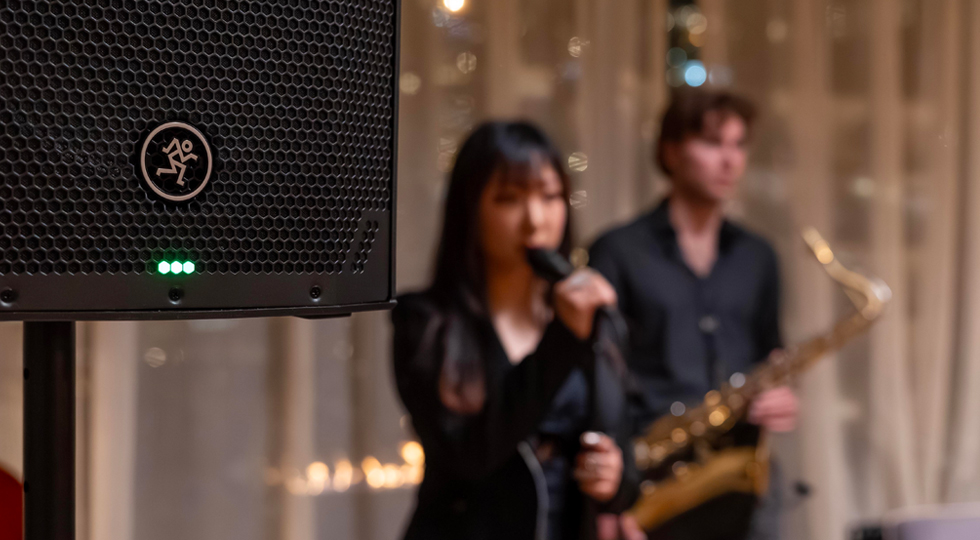 Singer holding microphone with saxophonist in background and Thumpv4 loudspeaker in foreground.