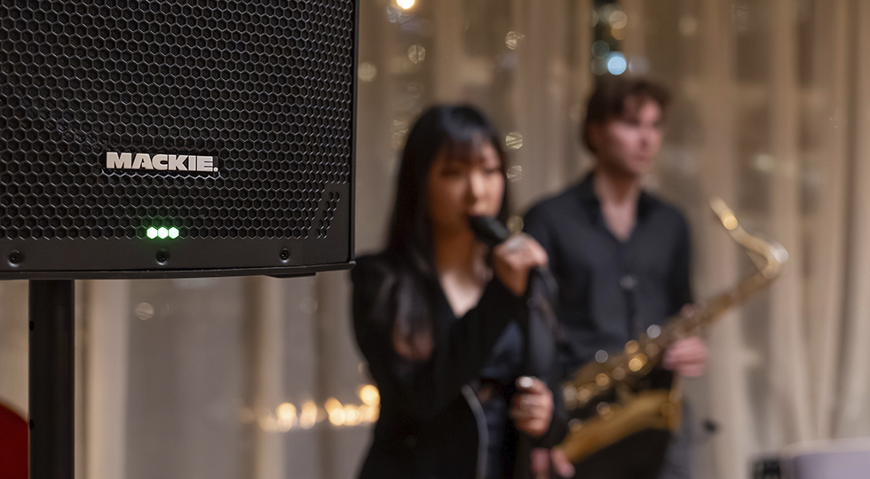 Singer holding microphone with saxophonist in background and Thumpv4 loudspeaker in foreground.